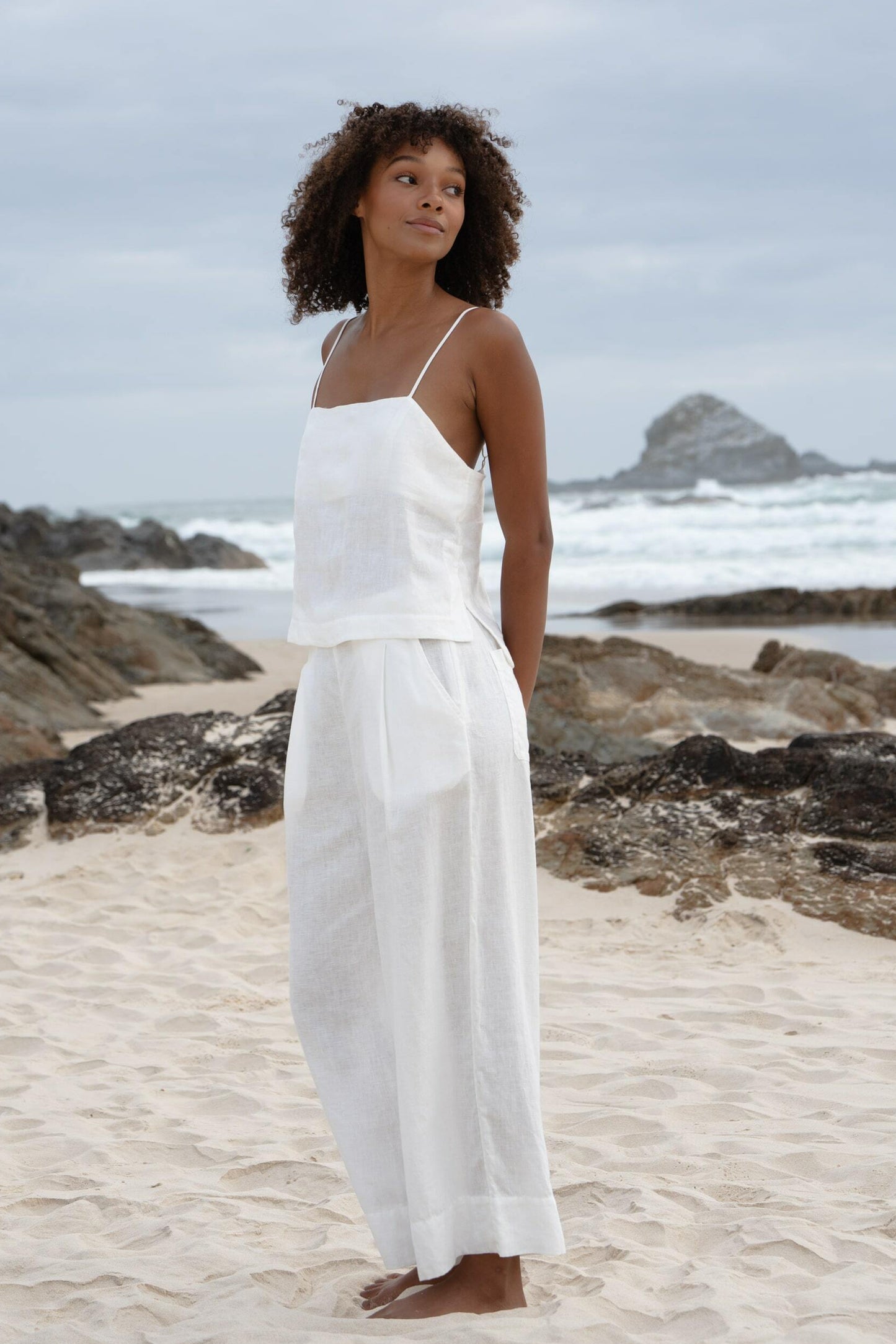 Ivy Linen Crop Pants in Ivory - side view showing front pleat and wide-leg linen crop silhouette, woman wearing matching ivory linen cami, sandy beach with coastal rocks backdrop - Lilly Pilly Collection