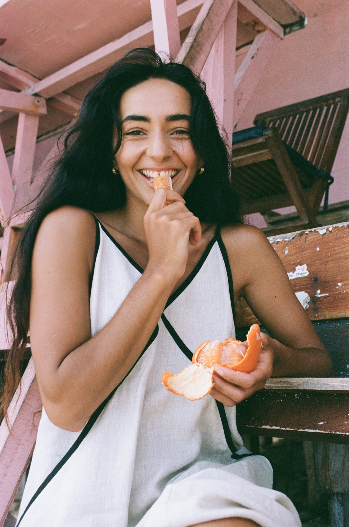 Azalea Linen Maxi Dress in Oatmeal - woman eating an orange, relaxed lifestyle shot, contrast black trim - Lilly Pilly Collection