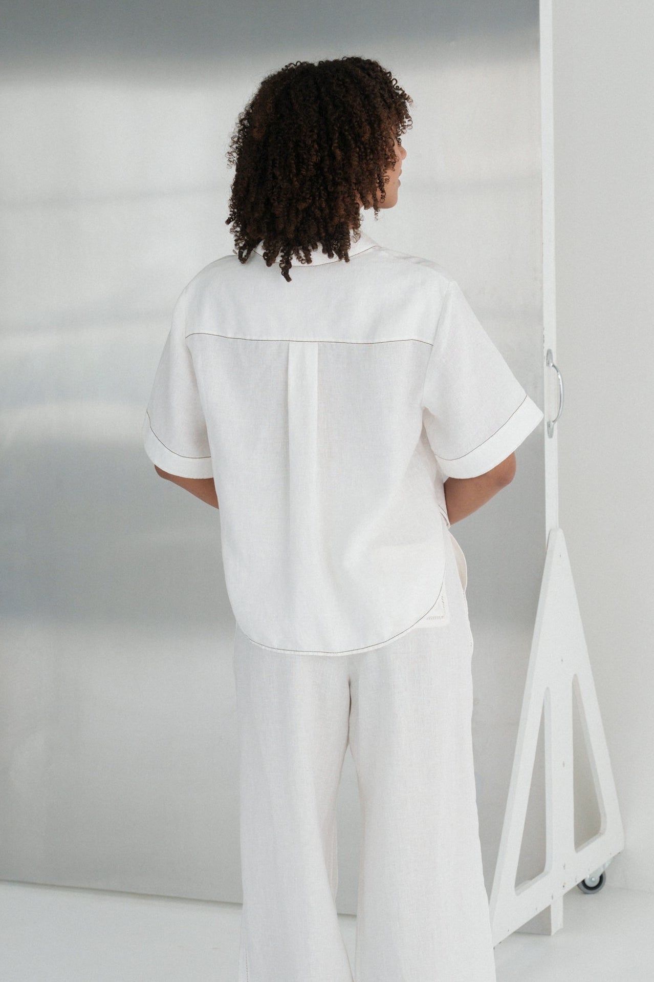 Back view of LILLY PILLY woman wearing an ivory Nika linen shirt top and Ivy linen pants standing in a minimalistic room with a mirror.