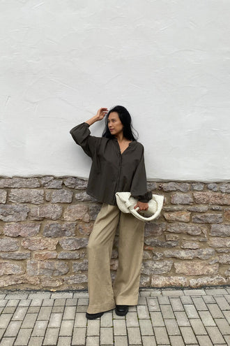 Ember Cotton Voile Shirt in Khaki - full length, holding woven clutch bag, standing against stone wall - Lilly Pilly Collection