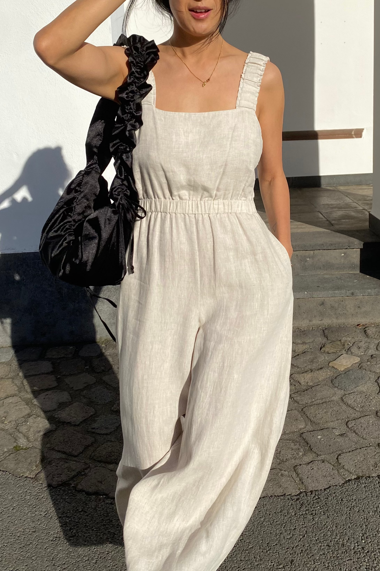 Front view of a woman wearing the Keely Linen jumpsuit in Oatmeal with  black shoulder bag- Lilly Pilly Collection