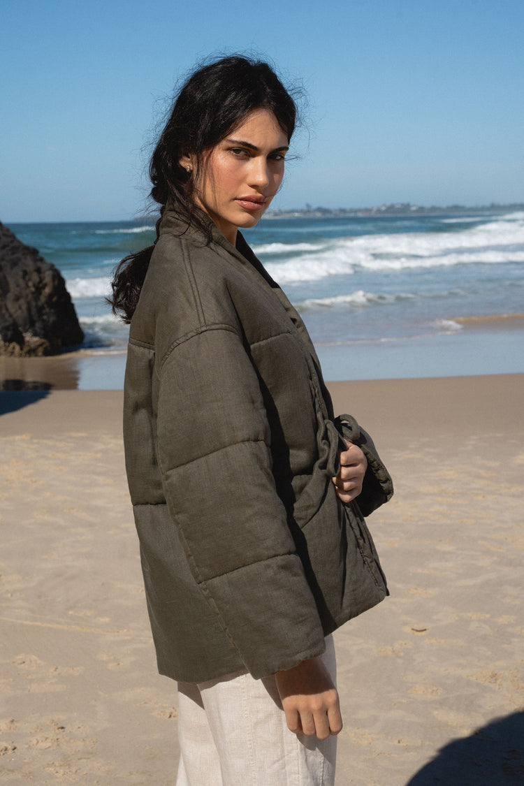 Aster Linen Coat in Khaki - side profile, tie-front and shawl collar, holding bag, worn over white trousers, standing on beach with ocean and rocks backdrop - Lilly Pilly Collection