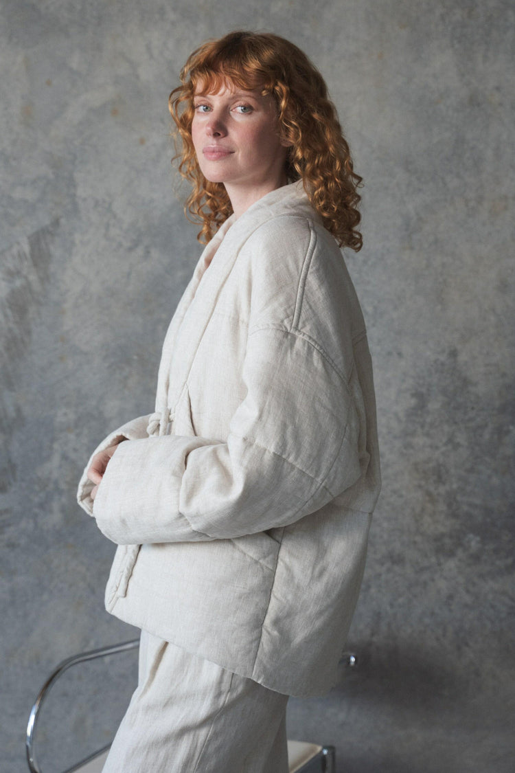 Aster Linen Coat in Oatmeal - side profile, arms crossed, worn over cream trousers, studio setting - Lilly Pilly Collection