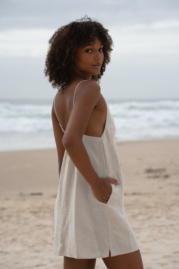Liz Linen Mini Slip Dress in Oatmeal - side profile, hand in pocket, adjustable strap detail, standing on beach with ocean backdrop - Lilly Pilly Collection