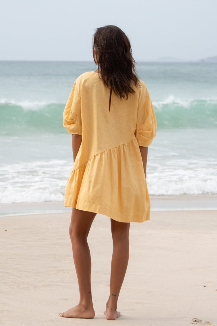 Layla Linen Short Dress in Butter Yellow - back view, keyhole button detail, asymmetric drop-waist hem, standing on beach with ocean backdrop - Lilly Pilly Collection