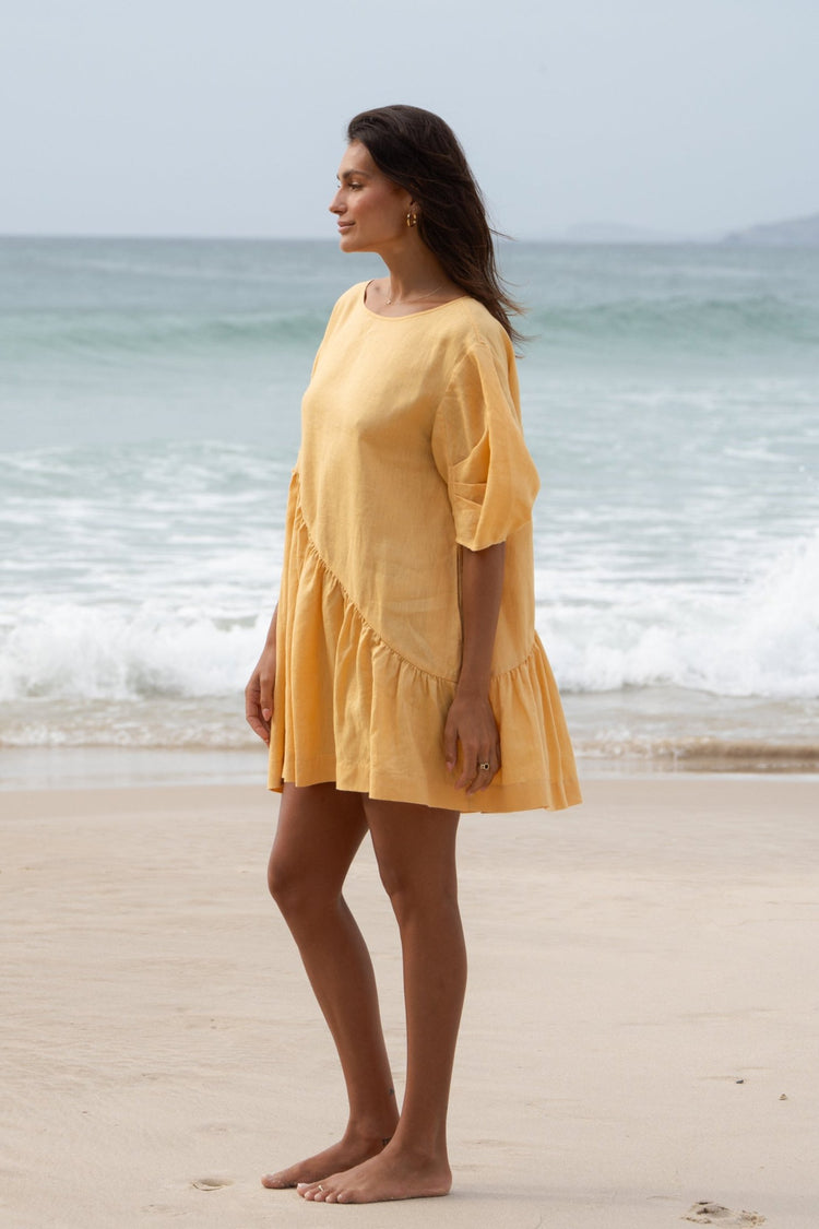 Layla Linen Short Dress in Butter Yellow - side profile, round neckline and puff sleeves, standing on beach with ocean backdrop - Lilly Pilly Collection