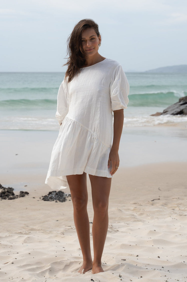 Layla Linen Mini Dress in Ivory - full length front view, round neckline and puff sleeves, asymmetric drop-waist hem, standing on beach with ocean backdrop - Lilly Pilly Collection