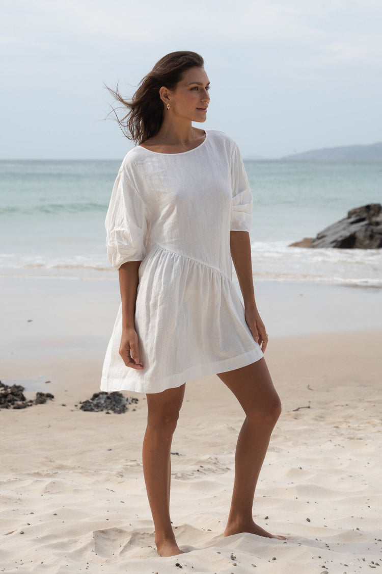 Layla Linen Mini Dress in Ivory - full length three-quarter view, round neckline and puff sleeves, standing on beach with ocean backdrop - Lilly Pilly Collection