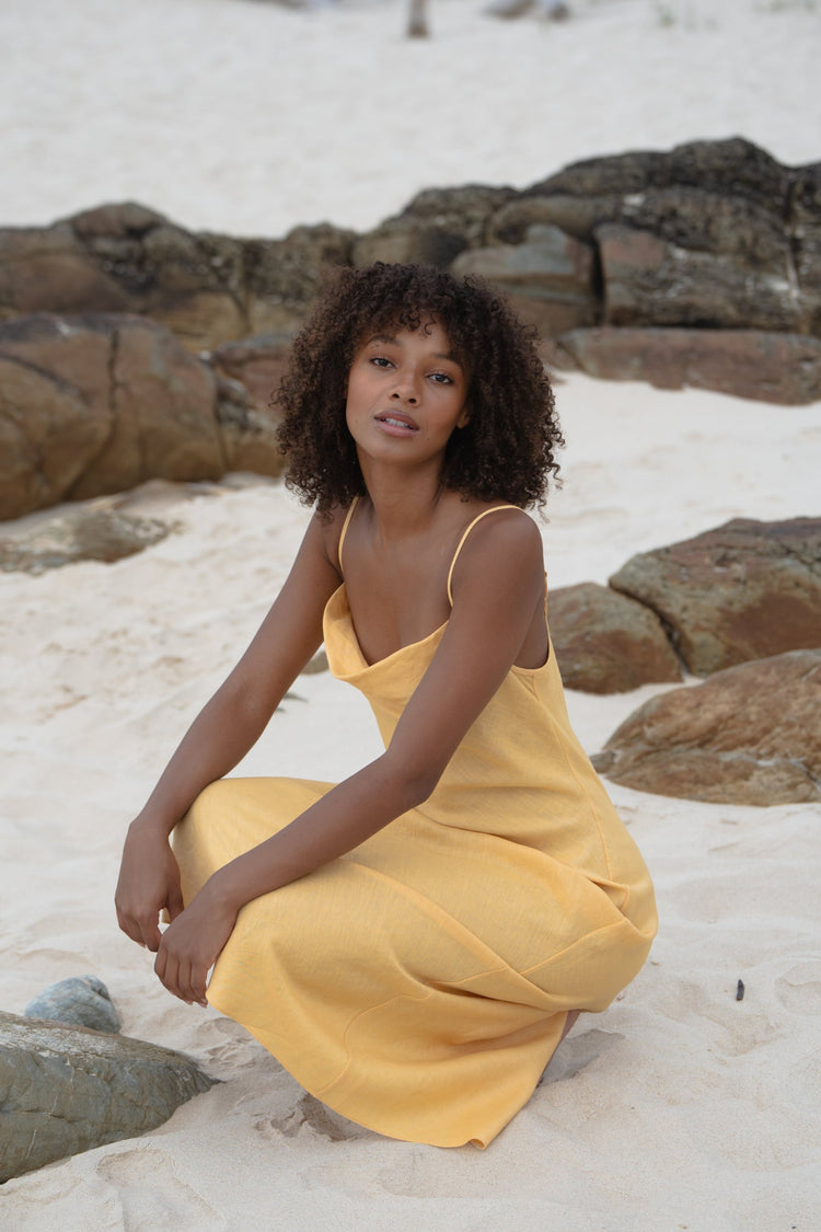 Allegria Bias Linen Maxi Dress in Butter Yellow - crouching on beach rocks, cowl neckline, sandy beach backdrop - Lilly Pilly Collection