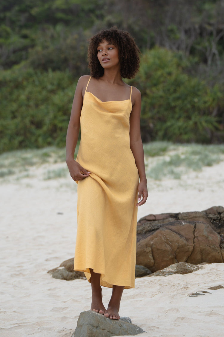 Allegria Bias Linen Maxi Dress in Butter Yellow - full length front view, cowl neckline and spaghetti straps, standing on a rock on sandy beach with coastal scrub backdrop - Lilly Pilly Collection