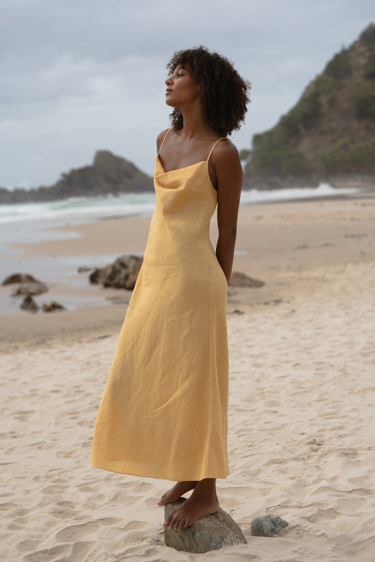 Allegria Bias Linen Maxi Dress in Butter Yellow - side profile, cowl neckline, standing on beach with ocean and headland backdrop - Lilly Pilly Collection
