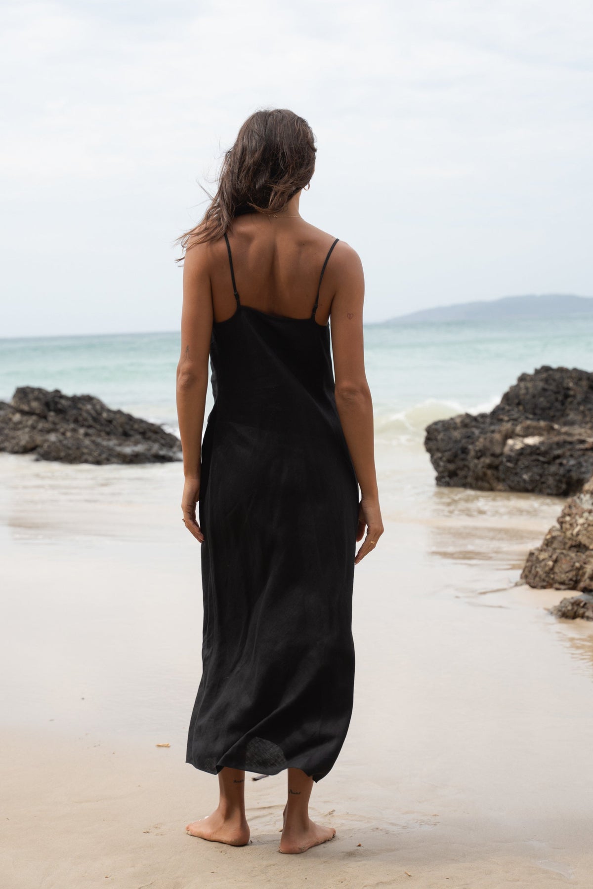 Back view of woman in a black Allegria linen dress standing on a beach with ocean view