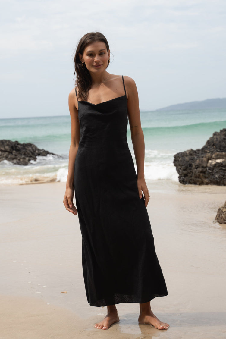 Allegria Bias Linen Maxi Dress in Black - front view, standing on a beach, ocean and rocks in the background - Lilly Pilly Collection