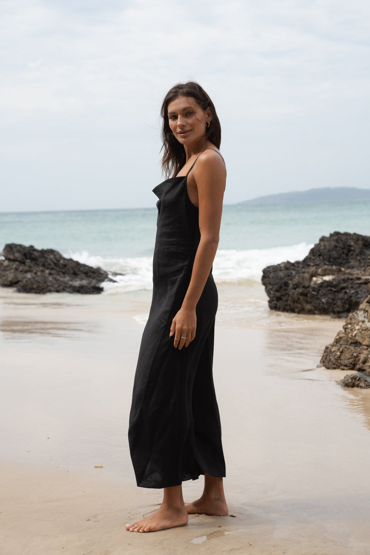 Allegria Bias Linen Maxi Dress in Black - side view, standing on a beach, ocean and rocks in the background - Lilly Pilly Collection