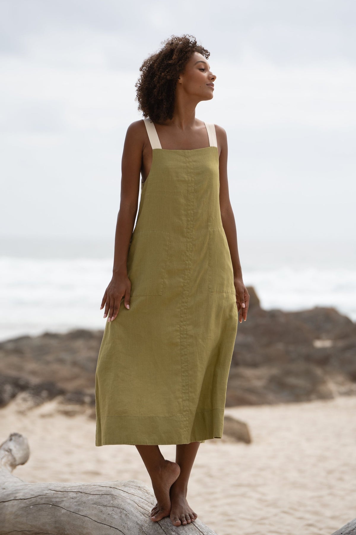 A woman standing on a log at the beach in an olive green Remi linen dress with adjustable white shoulder straps.