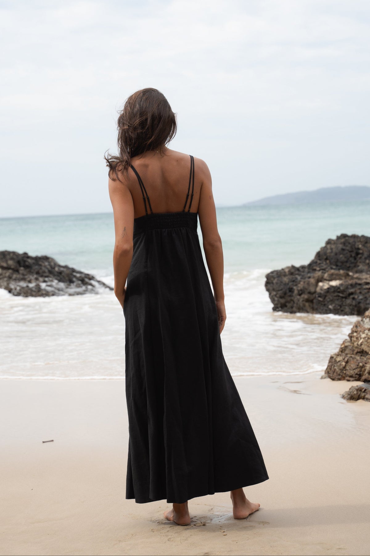 Back view of woman standing on a beach wearing a black Tori linen maxi dress with double adjustable shoulder straps and a curved empire neckline.