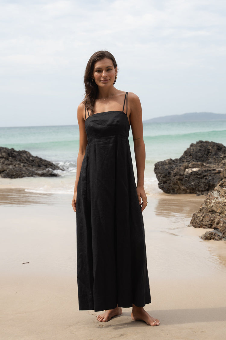 Tori Linen Midi Dress in Black - front view, double adjustable shoulder straps and curved empire neckline, standing on a beach - Lilly Pilly Collection