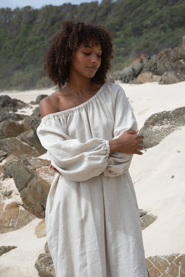 Lucia Linen Midi Dress in Oatmeal - front view, arms crossed, off-shoulder neckline and balloon sleeves, standing on rocky beach - Lilly Pilly Collection