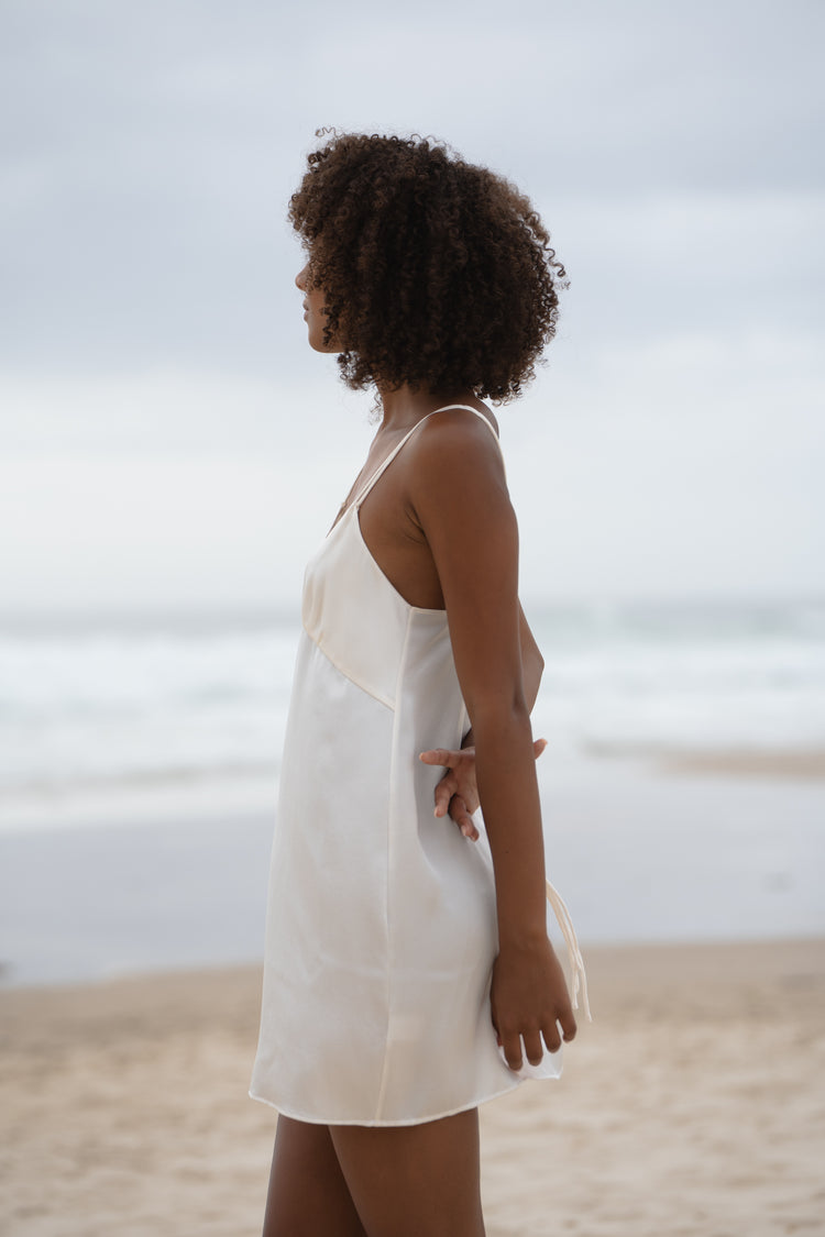 Ella Silk Mini Slip Dress in Ivory - side profile, hands behind back, standing on beach with ocean backdrop - Lilly Pilly Collection