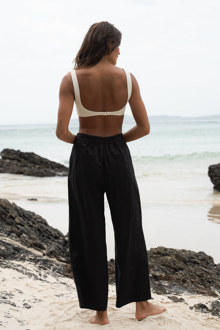 Ivy Linen Crop Pants in Black - back view showing elasticated waistband and wide-leg linen silhouette, woman wearing ivory square-neck crop bra top, coastal rock beach backdrop - Lilly Pilly Collection