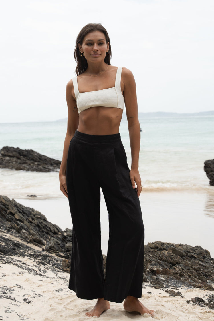 Ivy Linen Crop Pants in Black - full length front view, woman wearing Black wide-leg linen crop pants with ivory square-neck crop bra top, coastal rock beach backdrop - Lilly Pilly Collection