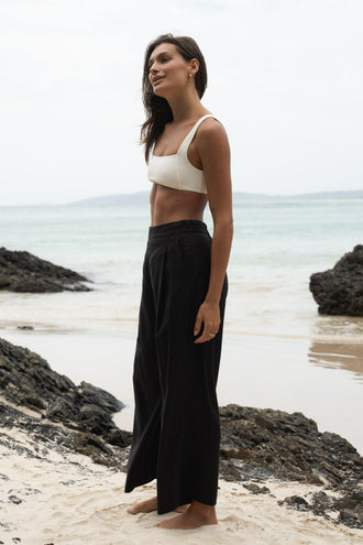Ivy Linen Crop Pants in Black - side view showing front pleat and wide-leg linen crop silhouette, woman wearing ivory square-neck crop bra top, coastal rock beach backdrop - Lilly Pilly Collection
