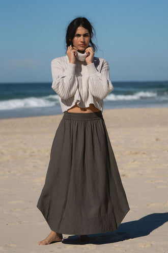 Hannah Linen Maxi Skirt in Khaki - front view, elasticated waist, worn with turtleneck knit, standing on beach with ocean backdrop - Lilly Pilly Collection
