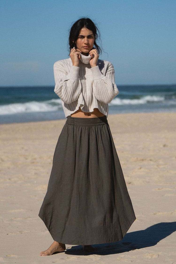 Hannah Linen Maxi Skirt in Khaki - front view, elasticated waist, worn with turtleneck knit, standing on beach with ocean backdrop - Lilly Pilly Collection