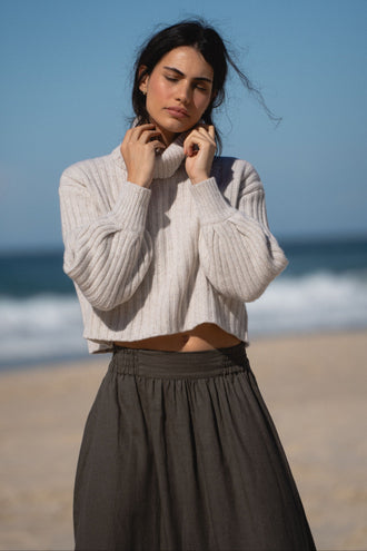 Hannah Linen Maxi Skirt in Khaki - close-up front view, worn with turtleneck knit, standing on beach with ocean backdrop - Lilly Pilly Collection