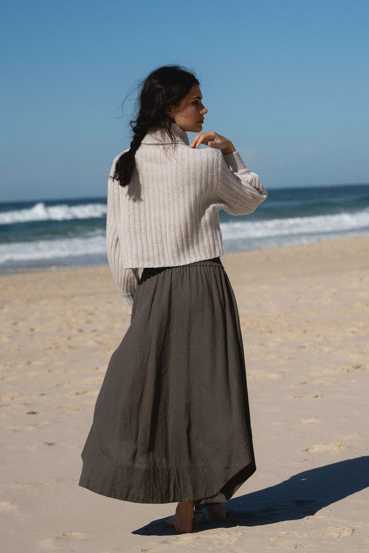 Hannah Linen Maxi Skirt in Khaki - back view, elasticated waist, worn with turtleneck knit, standing on beach with ocean backdrop - Lilly Pilly Collection