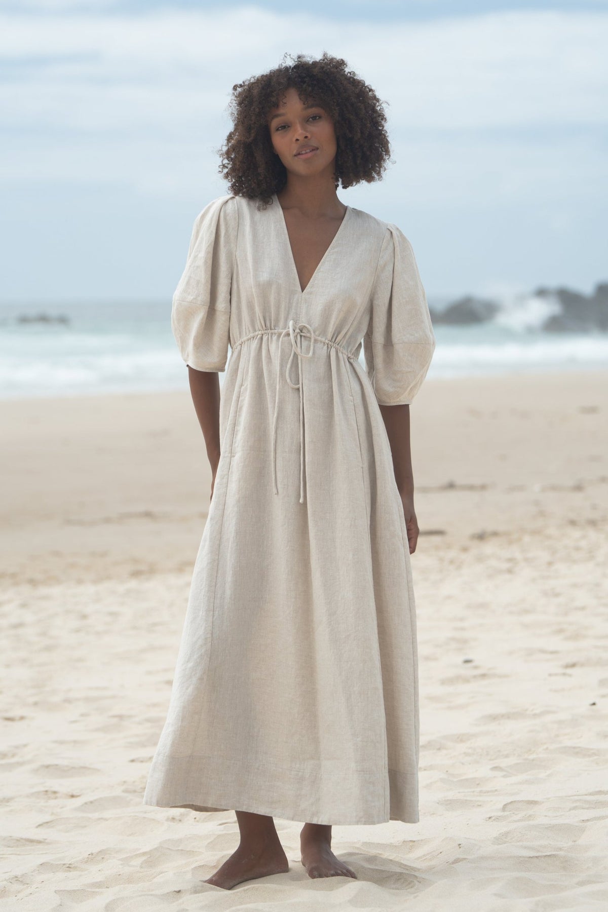 Front view of a woman wearing the Vida Linen maxi sleeve dress in Oatmeal on a beach - Lilly Pilly Collection