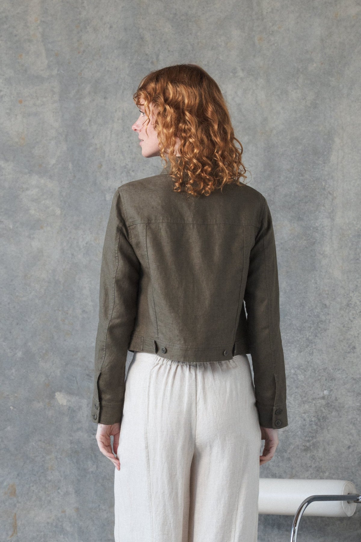 Back view of woman modelling a MILLY khaki linen jacket with denim-inspired details with a grey background