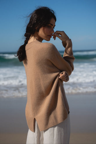 June Cashmere Knit in Camel Brown - back view, split hem detail, worn with Oli Linen Pants in Oatmeal, standing on beach with ocean backdrop - Lilly Pilly Collection
