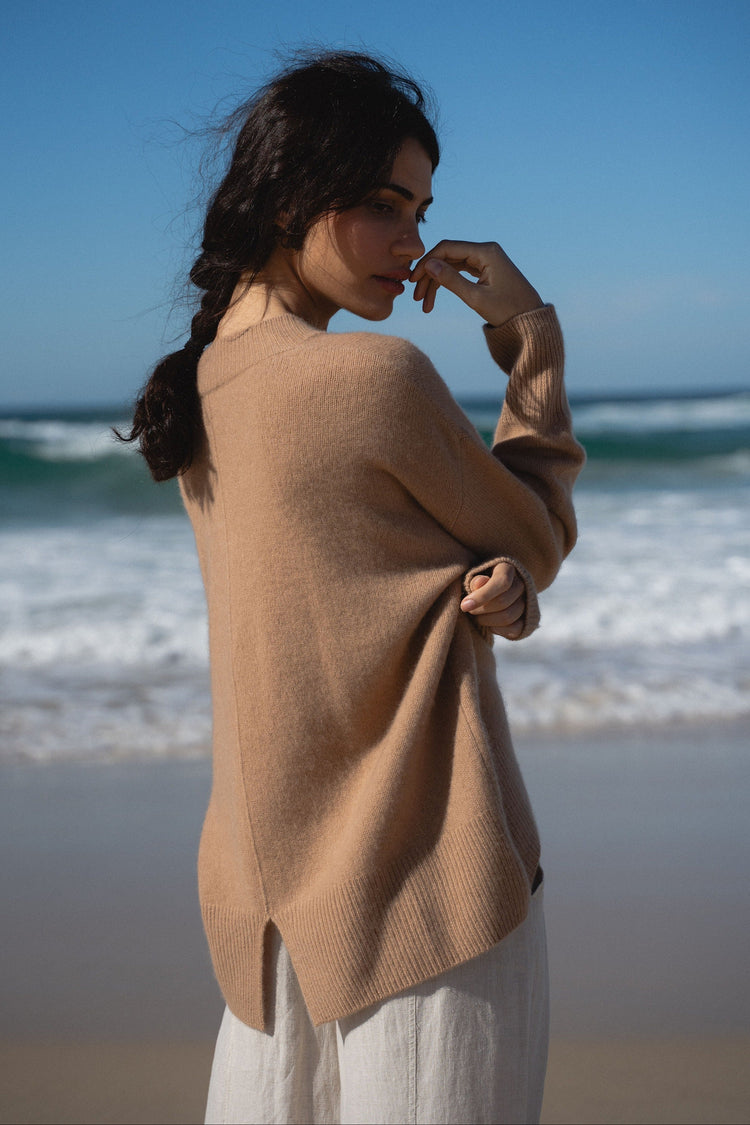 June Cashmere Knit in Camel Brown - back view, split hem detail, worn with wide-leg trousers, standing on beach with ocean backdrop - Lilly Pilly Collection