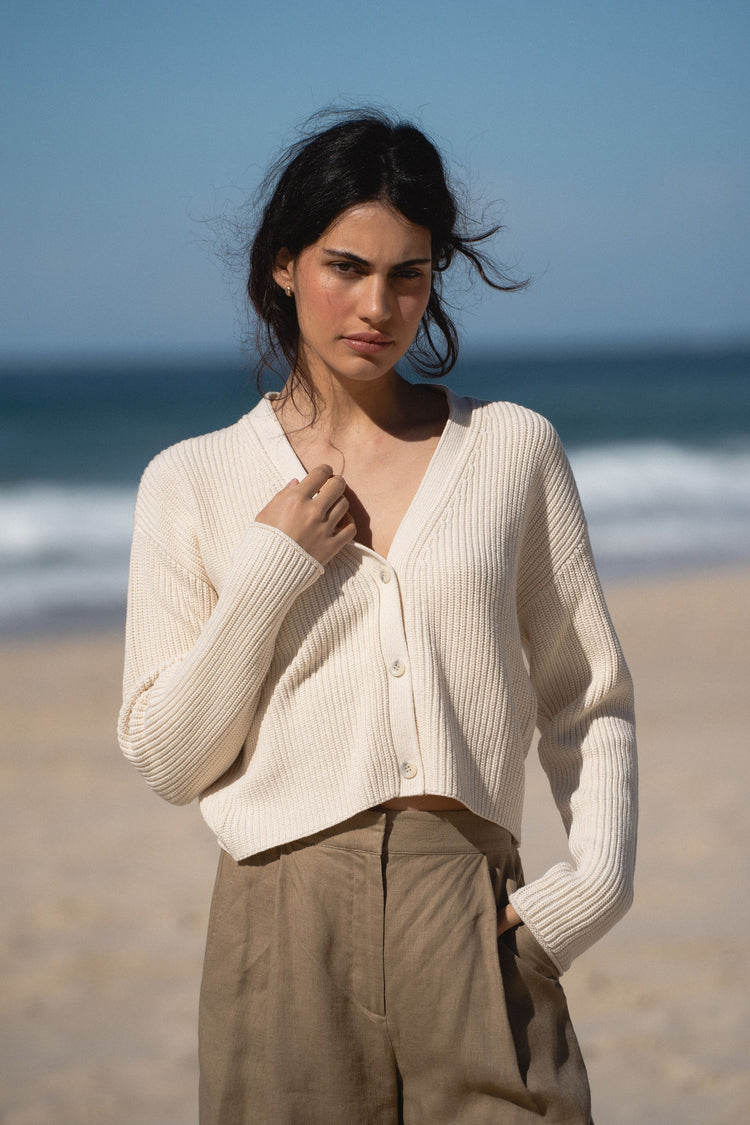 Bailey Cotton Cardigan in Cream - front view, V-neckline and button-front, worn open, holding collar, standing on beach with ocean backdrop - Lilly Pilly Collection