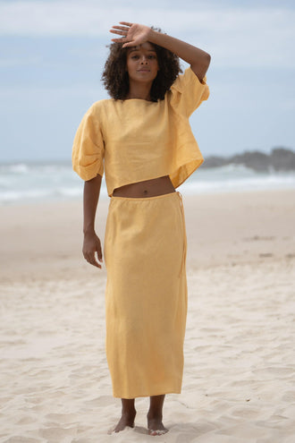Riley Bias Linen Midi Skirt in Butter - full length front view, woman wearing matching Butter linen puff-sleeve crop top, beach backdrop - Lilly Pilly Collection