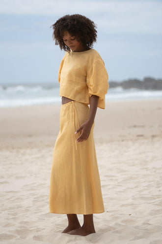 Riley Bias Linen Midi Skirt in Butter - side view, woman wearing matching Butter linen puff-sleeve crop top, beach backdrop - Lilly Pilly Collection