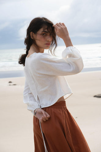 Louisa Linen Top in Ivory - side view, arm raised, boat neckline and balloon sleeves, worn with rust skirt, standing on beach with ocean backdrop - Lilly Pilly Collection