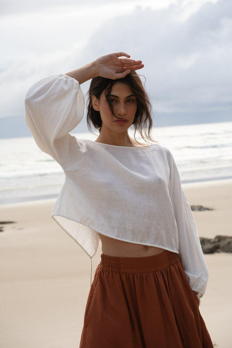 Louisa Linen Top in Ivory - front view, hand shielding eyes, boat neckline and balloon sleeves, worn with rust skirt, standing on beach with ocean backdrop - Lilly Pilly Collection