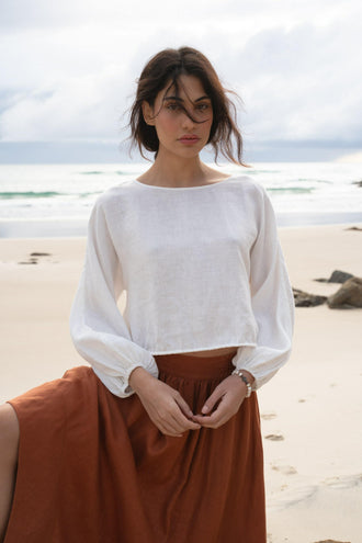 Louisa Linen Top in Ivory - front view, boat neckline and balloon sleeves, worn with rust skirt, standing on beach with ocean backdrop - Lilly Pilly Collection