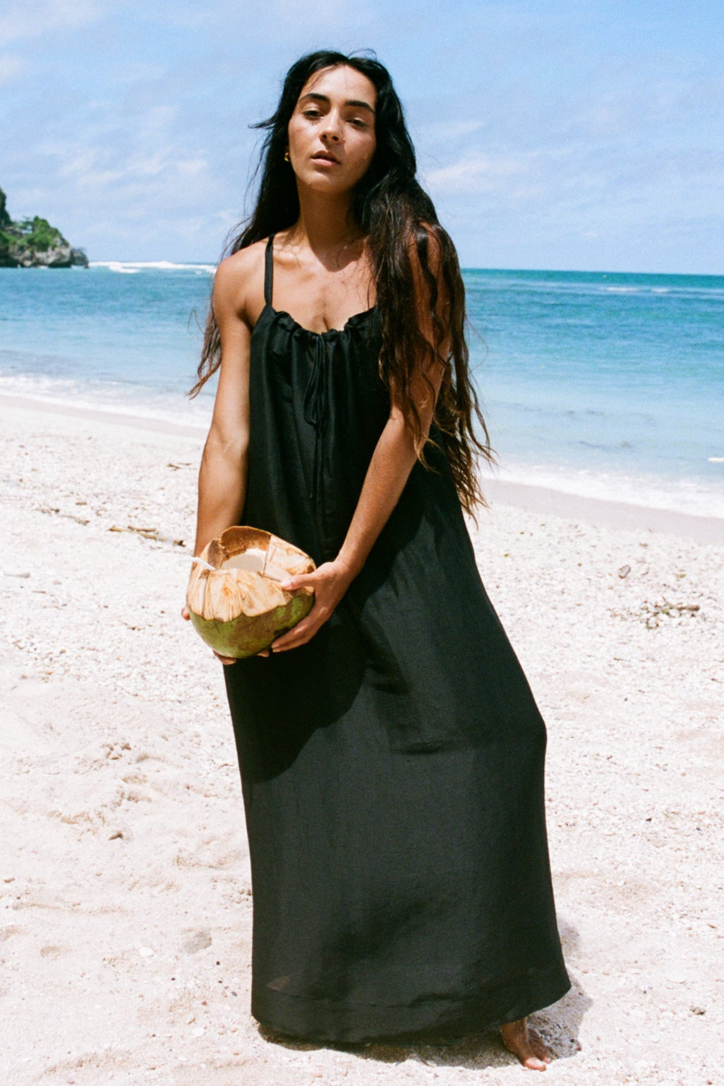 Woman in a black dress holding a coconut on a beach with ocean and sky in the background