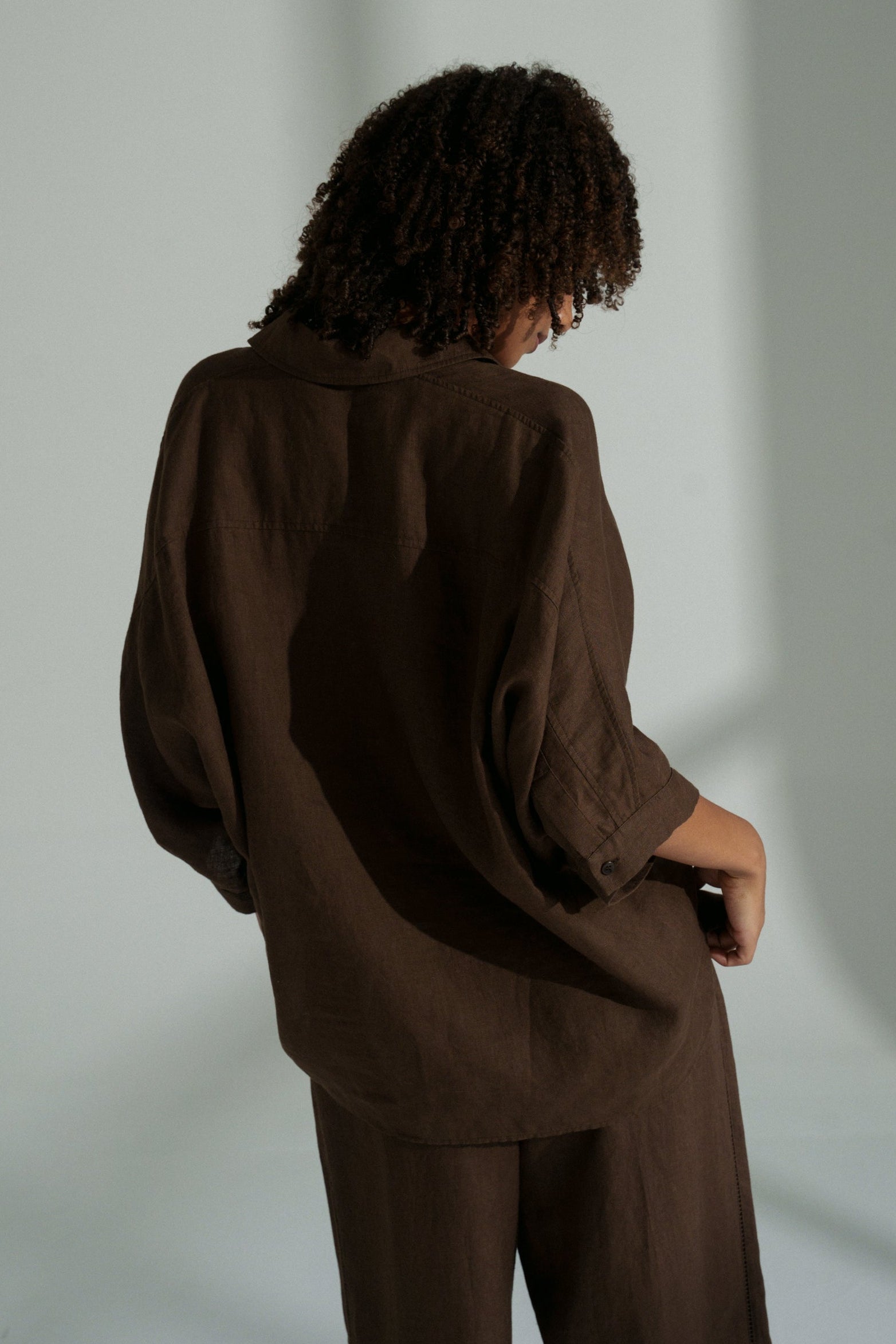 LILLY PILLY woman wearing a chocolate brown Pixie linen shirt and linen pants set showing the back view while against a plain background