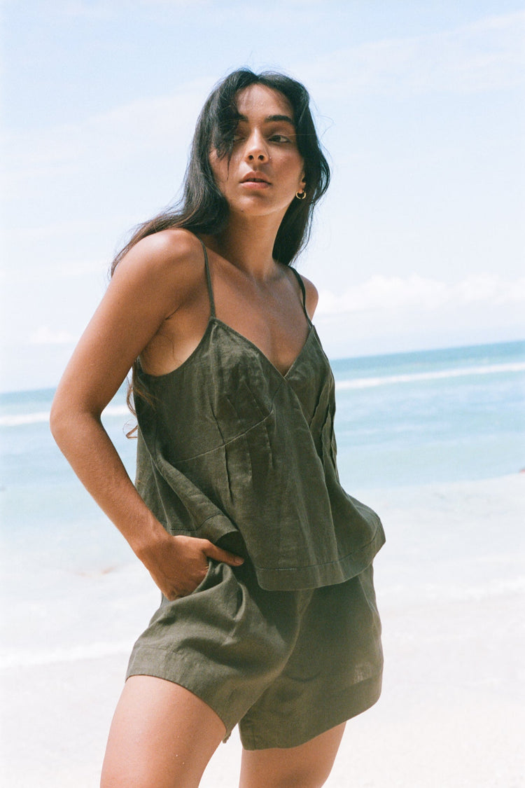 Bea Linen Cami in Khaki - three-quarter view, hand in pocket, V-neckline and pintuck detail, styled with matching Gaia Linen Shorts, beach and ocean backdrop - Lilly Pilly Collection