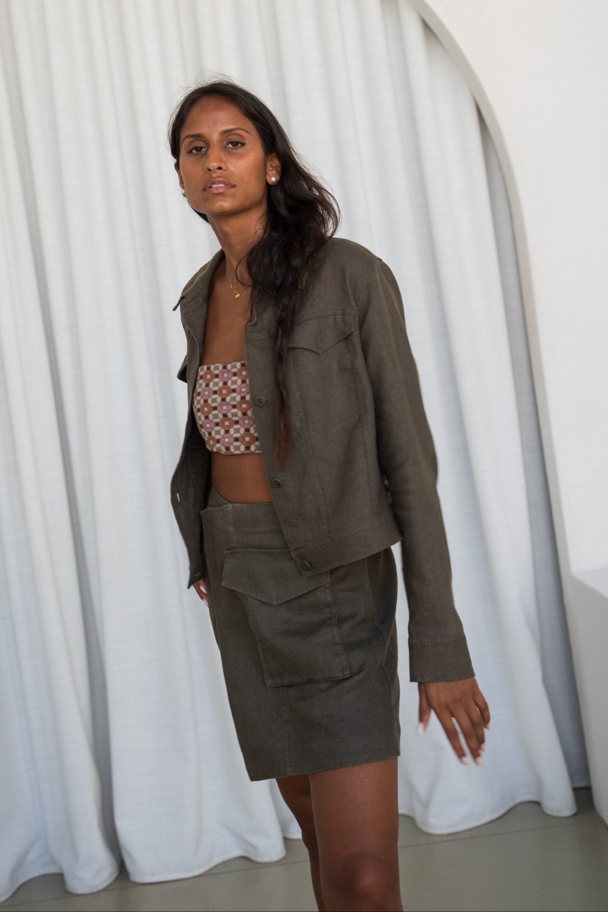 Side view of woman wearing a khaki green Milly linen jacket and Betty linen skirt set against a white curtain background
