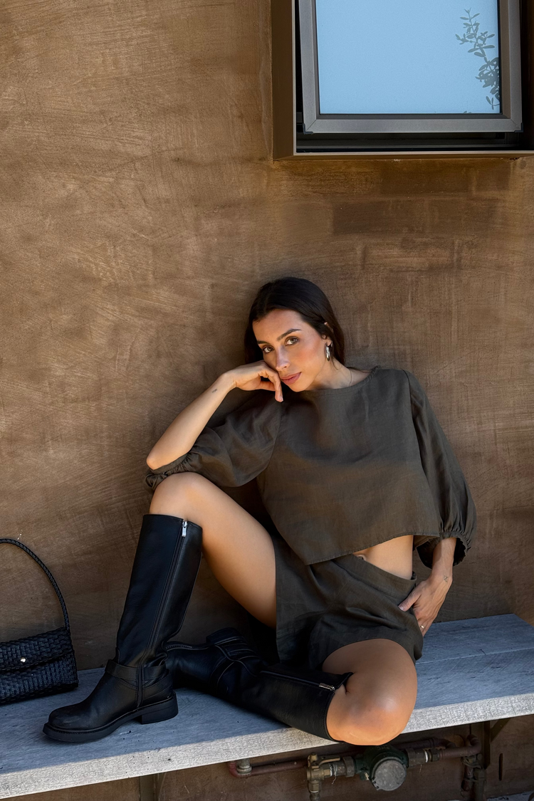 Demi Linen Top in Khaki - woman seated, hand to chin, round neckline and puff sleeves, styled with matching linen shorts, textured wall backdrop - Lilly Pilly Collection