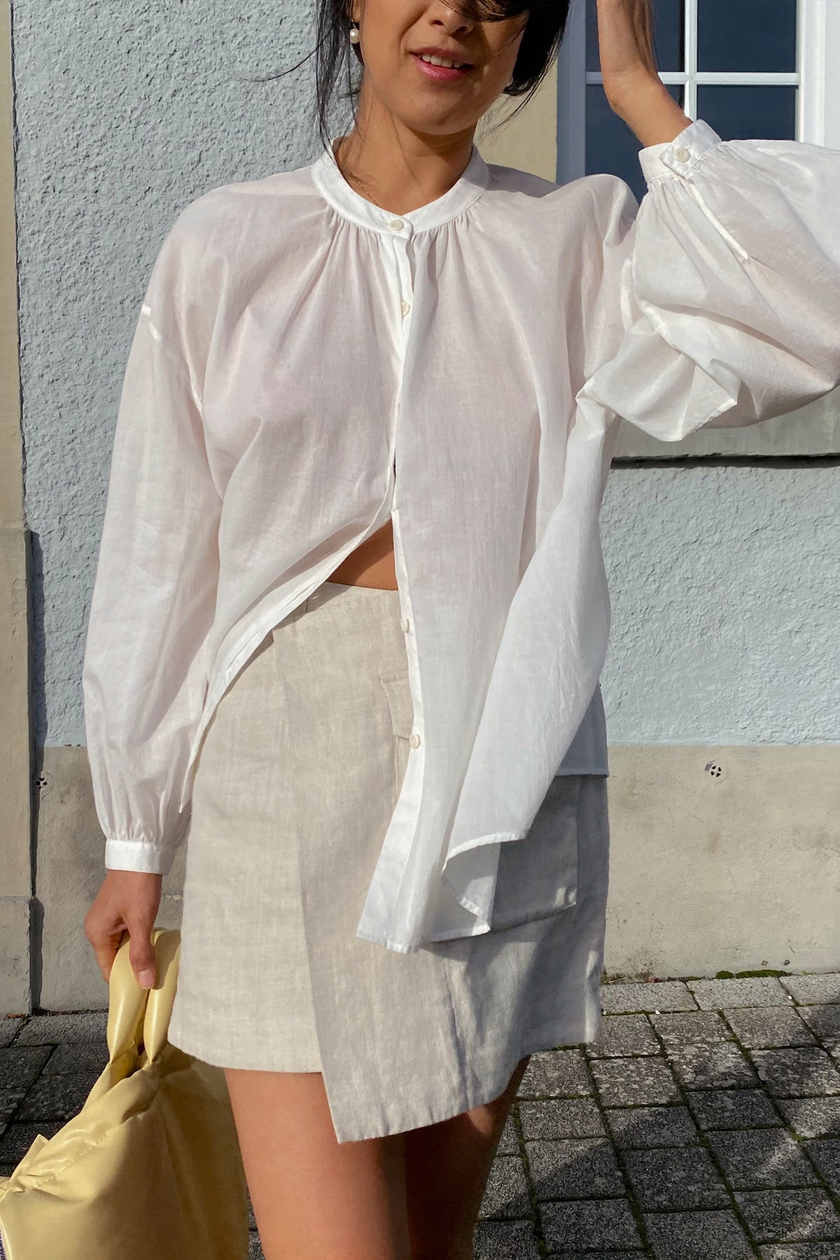LILLY PILLY woman in an ivory white Ember cotton voile blouse and beige shorts standing against a light blue wall.