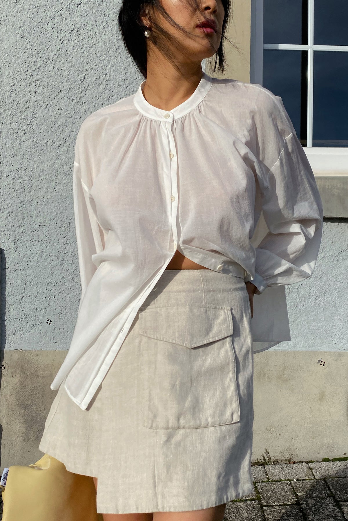 LILLY PILLY woman in an ivory white Ember cotton voile top and beige skirt against a gray wall.