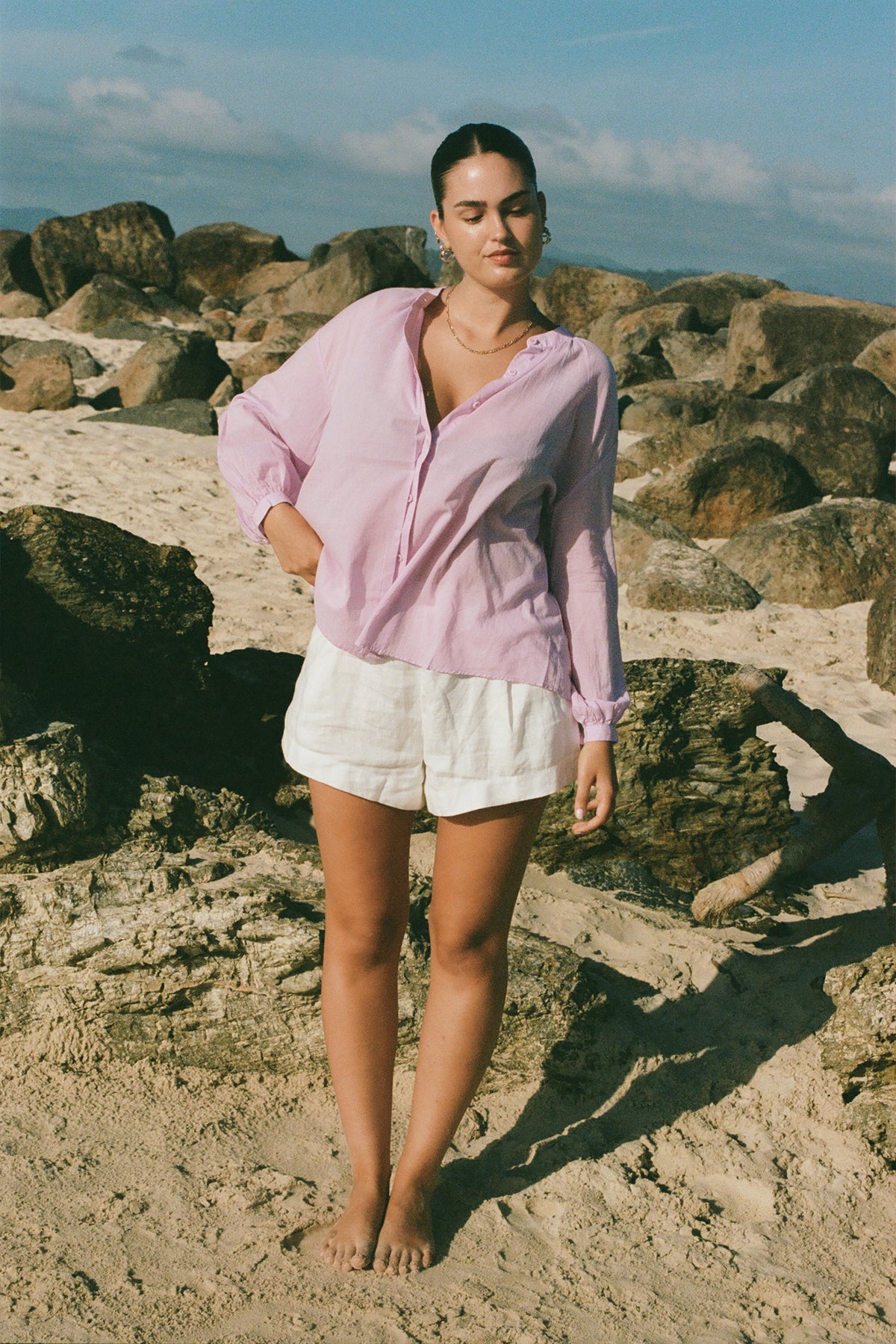 LILLY PILLY woman in an orchid Ember cotton voile top and white shorts  standing on a rocky beach.