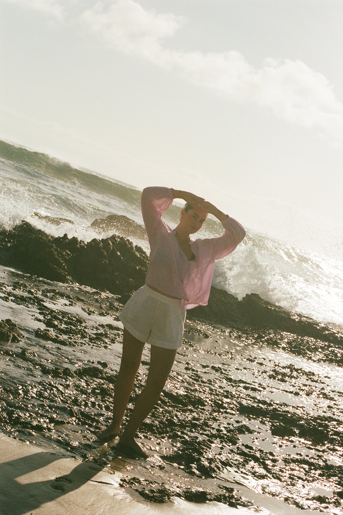 LILLY PILLY woman in an orchid Ember cotton voile top and white shorts  standing on a rocky beach with waves crashing in the background