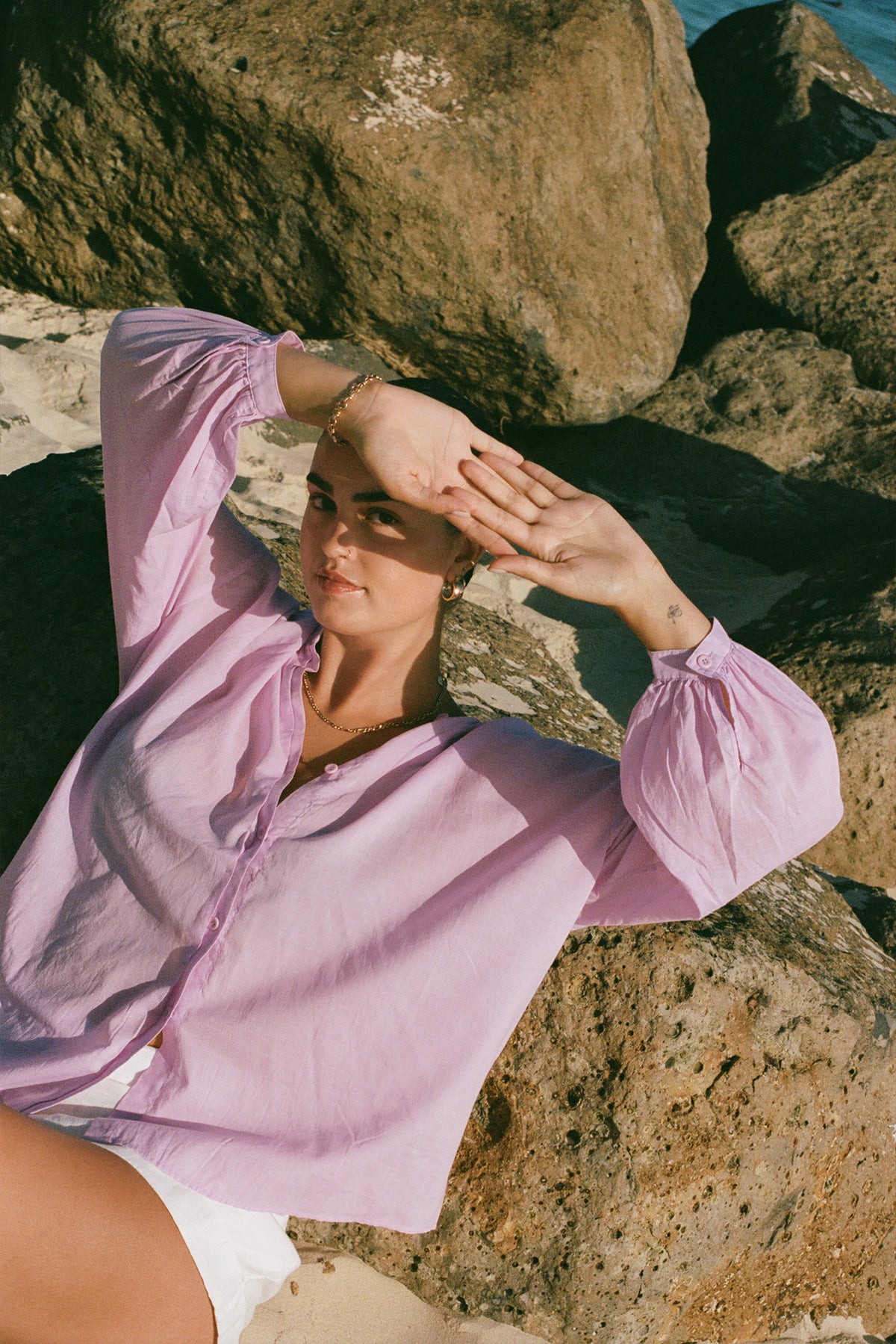 LILLY PILLY woman in an orchid Ember cotton voile top and white shorts sitting on rocks by the water